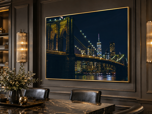 A large framed wall art print depicting the Brooklyn Bridge at night over a city skyline, displayed in a dark, elegant dining room setting with a marble table and leather chairs.