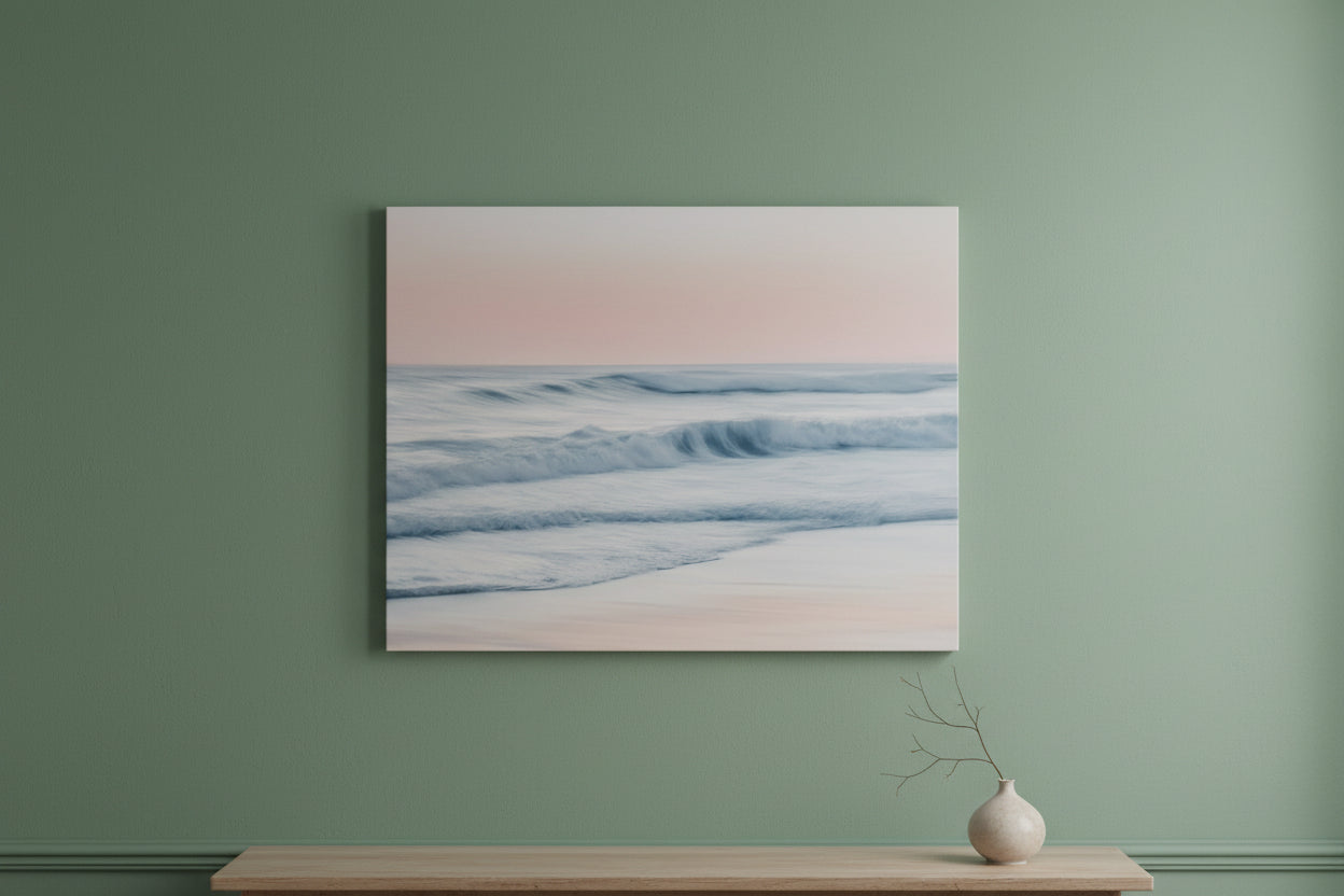 A framed wall art print showing a soft seascape with pale pink sky, blue-gray waves, and white foam, displayed on a sage green wall above a light wood console with a small vase.
