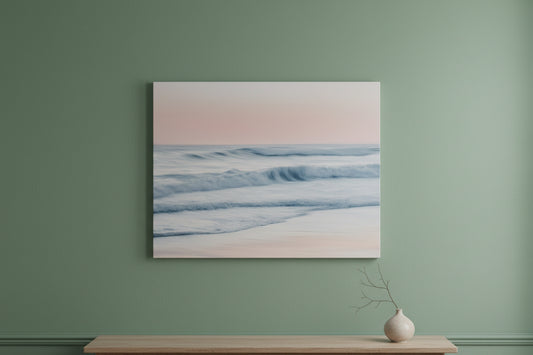 A framed wall art print showing a soft seascape with pale pink sky, blue-gray waves, and white foam, displayed on a sage green wall above a light wood console with a small vase.