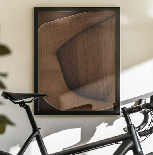 Caramel lines I by Alma, black-framed artwork above a bicycle; a folded caramel-brown ribbon-like form with fine grain and sweeping curves.