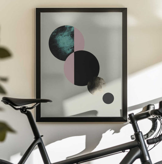 Celestial Circles by Emanuela Carratoni, black-framed artwork above a bicycle; split teal, pink and black circles descending toward smaller gray discs.